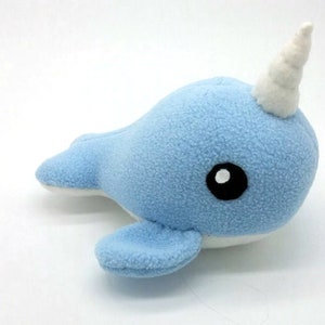 May include: A light blue plush narwhal toy with a white curled horn and black eyes. The toy is made of soft, fuzzy fabric.