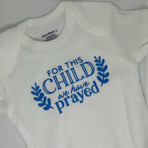 May include: White baby onesie with blue text that reads "FOR THIS CHILD we have prayed." The text is surrounded by blue leaf designs. The onesie is a newborn size.