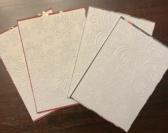 Könnte beinhalten: Eine Sammlung weißer Weihnachtskarten mit geprägten Schneeflocken- und Wirbelmustern. Die Karten haben eine strukturierte Oberfläche und sind auf einer dunklen Holzoberfläche angeordnet. Einige Karten haben rote Ränder.