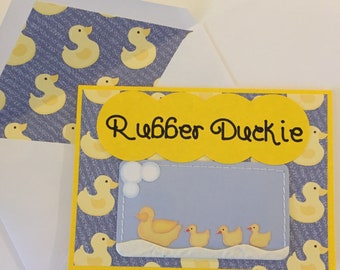 May include: A yellow card with a blue background and a yellow rubber duckie design. The card says "Rubber Duckie" in black lettering. There are three yellow rubber duckies in a row on the card.