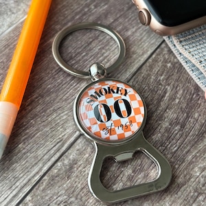 May include: A silver bottle opener keychain with a round, checkered orange and white design. The design features the word "SMOKEY", the numbers "00", and the text "est. 1953". The keychain has a silver ring.