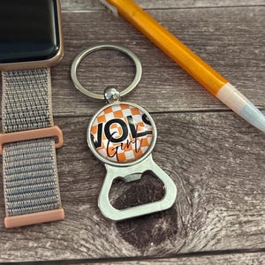 Może przedstawiać: Srebrny otwieracz do butelek z brelokiem z okrągłym wzorem w kratkę w kolorze pomarańczowym i białym. Wzór zawiera napis "VOLS Girl" w kolorze czarnym.