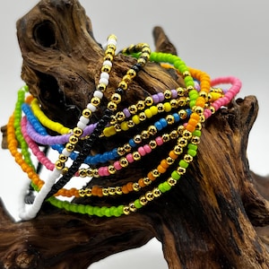 May include: A collection of colorful beaded bracelets in various shades, including pink, green, blue, and yellow. Each bracelet features small, vibrant beads and gold-colored accents. The bracelets are displayed on a piece of textured wood.
