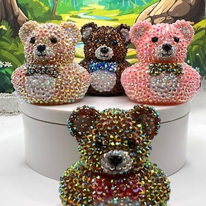 May include: Four jeweled teddy bear figurines in shades of pink, brown, and gold. Each bear has a bow tie and is covered in iridescent rhinestones. The bears are arranged on a white surface with a forest-themed backdrop.