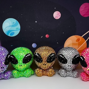 May include: Five small alien figurines made of rhinestones. The aliens are in different colors: purple, green, gold, silver, and pink.