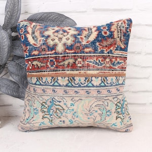 May include: Square decorative pillow with a vintage rug design. The pillow features a patchwork of patterns in blue, red, and beige. The pillow is placed in front of a white brick wall and a decorative plant.
