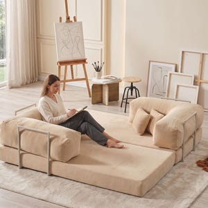 May include: A beige modular sofa with a corduroy texture and metal armrests. The sofa is arranged on a light beige rug. The room also features an easel with artwork, a side table, and a modern light fixture.
