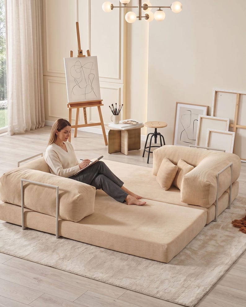 May include: A beige modular sofa with a corduroy texture and metal armrests. The sofa is arranged on a light beige rug. The room also features an easel with artwork, a side table, and a modern light fixture.