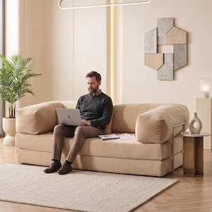 May include: A beige corduroy sofa with rounded arms. A person is seated on the sofa, using a laptop. The room features a neutral color palette, a rectangular light fixture, a decorative wall piece, and a small side table.