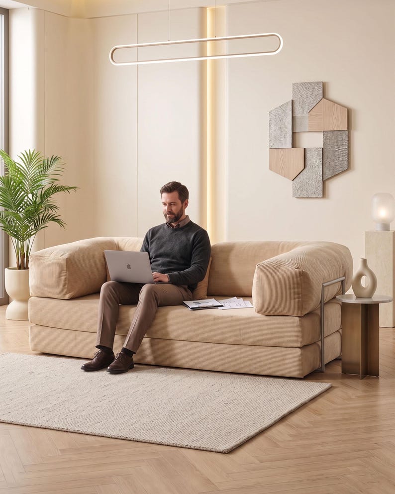 May include: A beige corduroy sofa with rounded arms. A person is seated on the sofa, using a laptop. The room features a neutral color palette, a rectangular light fixture, a decorative wall piece, and a small side table.