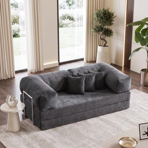 May include: A modern, gray convertible sofa with corduroy fabric and three matching throw pillows. The sofa has a low profile and a metal frame. It is placed on a light-colored rug in a well-lit room.