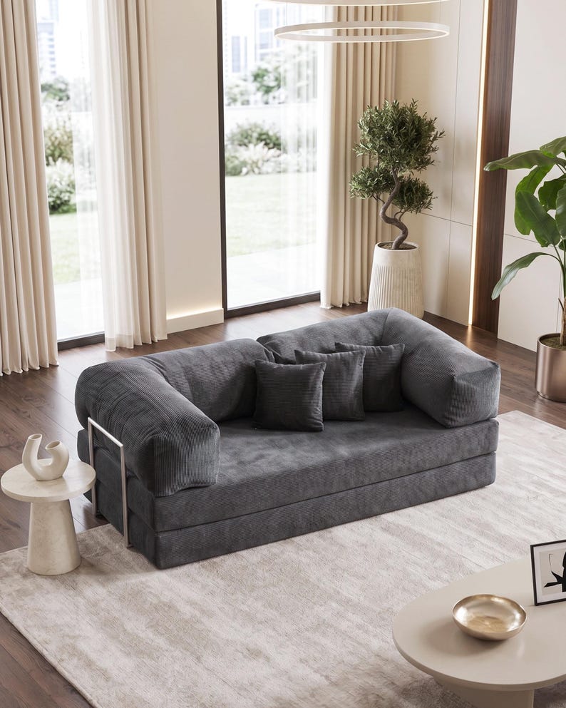 May include: A modern, gray convertible sofa with corduroy fabric and three matching throw pillows. The sofa has a low profile and a metal frame. It is placed on a light-colored rug in a well-lit room.
