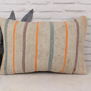 Puede incluir: Cojín rectangular beige con rayas verticales en naranja, gris azulado y marrón. El cojín está hecho de una tela tejida y se coloca sobre una superficie blanca con un fondo de pared de ladrillo.