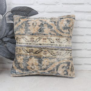 May include: A decorative throw pillow with a vintage, hand-woven design. The pillow features a beige and blue color scheme with intricate patterns and a subtle distressed look.