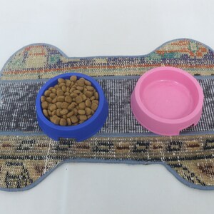 May include: A dog bowl set featuring a bone-shaped rug with a blue bowl filled with dog food and a pink bowl. The rug has a patterned design with blue trim. The bowls are made of plastic.