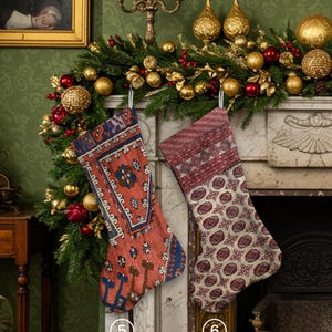 Pu&ograve; includere: Due calze di Natale sono appese a un caminetto decorato con ghirlande, ornamenti e candele. Una calza &egrave; rossa con un motivo blu e crema, l'altra &egrave; rossa con un motivo floreale color crema.