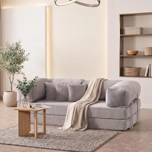May include: A modern living room setup featuring a light gray corduroy sofa with matching pillows and a beige throw blanket. A small wooden side table holds a vase of greenery and a magazine. A gray rug and a decorative light fixture complete the scene.