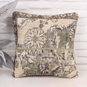 May include: Square decorative pillow with a vintage rug design. The pillow features a beige, brown, and green color palette with floral and geometric patterns. The pillow is approximately 16 inches square.