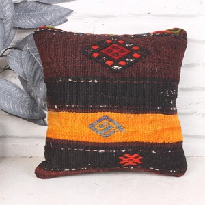 May include: A decorative square pillow with a geometric design. The pillow features a color block pattern with bands of burgundy, black, and orange. The pillow has diamond and geometric shapes in red, gray, and orange.