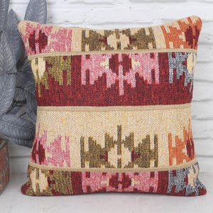 May include: Square decorative pillow with a woven design. The pillow features horizontal bands of red, beige, and patterned sections in pink, orange, and brown. The pillow is placed in front of a white brick wall.