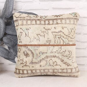 May include: Square beige pillow with a brown geometric pattern and a brown horizontal stripe. The pillow is placed in front of a white brick wall and next to a decorative plant.