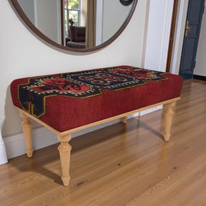 May include: A rectangular bench with a red fabric top featuring a geometric pattern in black, gold, and red. The bench has light-colored wooden legs. The bench is placed in front of a large, round mirror.