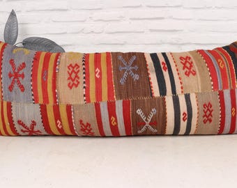 May include: A long, rectangular cushion with a patchwork design. The cushion is made from a variety of fabrics in shades of red, brown, beige, and white. The fabric is woven with intricate patterns and designs.