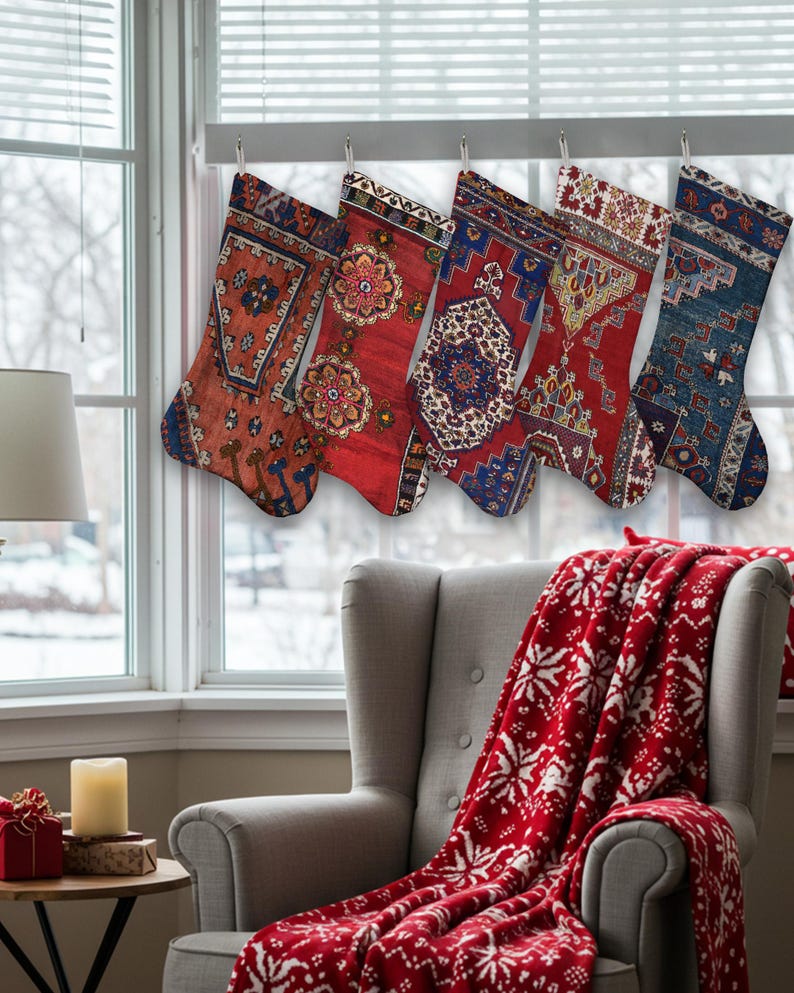 Pu&ograve; includere: Cinque calze natalizie con intricati motivi di tappeti colorati sono appese davanti a una finestra. Le calze presentano sfumature di rosso, blu e arancione. Una coperta rossa con disegni di fiocchi di neve bianchi &egrave; drappeggiata su una poltrona grigia.