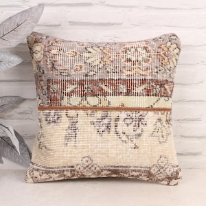 May include: Square decorative pillow with a patchwork design. The pillow features a mix of beige, brown, and muted colors, with a vintage rug-like pattern. The pillow is placed against a white brick wall.