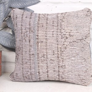 Può includere: Cuscino quadrato decorativo con un motivo tessuto e strutturato nei toni del grigio e del marrone. Il cuscino ha un dettaglio a righe verticali sul lato sinistro. Il cuscino è posto davanti a un muro di mattoni bianchi.