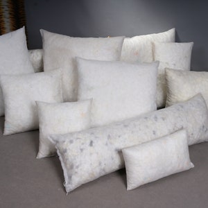 May include: A collection of white, fluffy pillows in various shapes and sizes. The assortment includes square, rectangular, and bolster pillows, all filled with a soft, white material. The pillows are arranged on a gray surface.