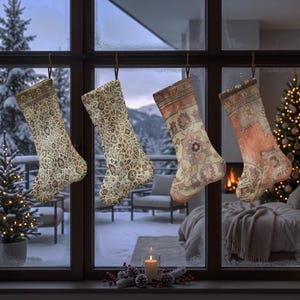 Op de afbeelding: Vier kerstsokken hangen op een rij voor een raam. De eerste twee sokken hebben ingewikkelde beige en bruine bloemmotieven. De derde sok toont een geometrisch medaillonontwerp in oranje en crème. De vierde is oranje en beige van kleur.