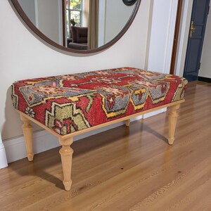 May include: A rectangular bench with a patterned red, yellow, and gray upholstered top. The bench has light-colored wooden legs. The bench is placed in front of a wall with a large round mirror.