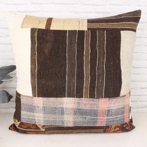 May include: A square decorative pillow with a patchwork design. The pillow features a combination of beige, brown, and cream-colored fabrics. The design includes vertical stripes, geometric patterns, and a mix of textures, creating a rustic and textured appearance.