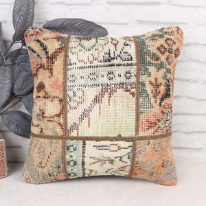 May include: Square decorative pillow with a patchwork design. The pillow features a mix of colors including beige, orange, and teal. The pillow is placed in front of a white brick wall and next to a decorative plant.