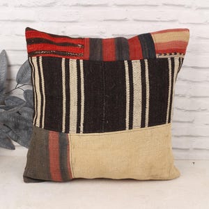 May include: Square cushion with a patchwork design. The top section features red and black stripes, the middle has vertical black and white stripes, and the bottom is a beige colour. The cushion is made of a woven fabric.