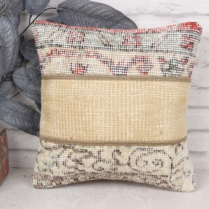 May include: Square decorative pillow with a beige center panel and patterned top and bottom sections. The top section has red, black, and white accents. The pillow is placed in front of a white brick wall.