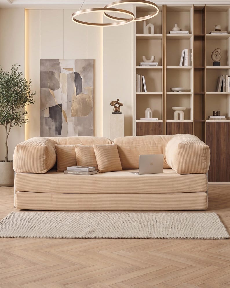 May include: A beige corduroy sofa with matching throw pillows and a laptop. The sofa sits on a light beige rug, with a wooden floor in the foreground. A modern living room setting with a neutral color palette.