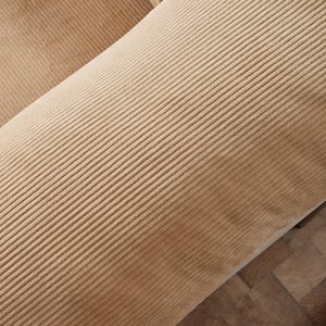 May include: Close-up of a section of a tan corduroy fabric. The fabric has a ribbed texture and is angled across the frame. The corduroy is a warm, neutral color, and the texture is clearly visible.
