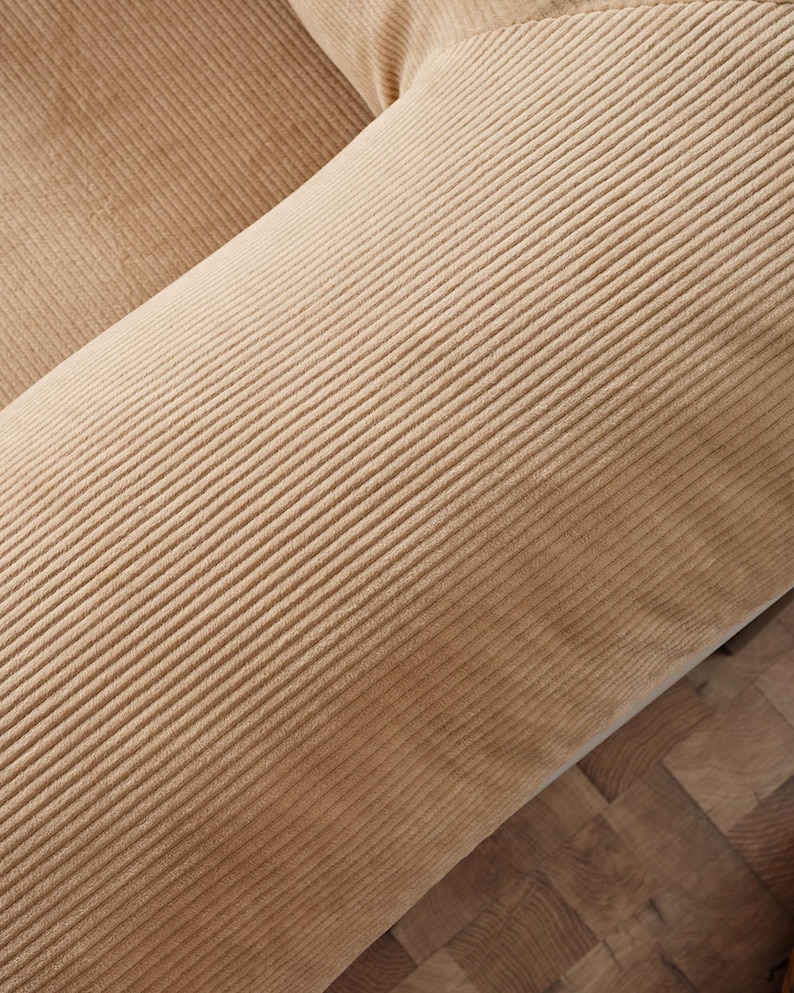 May include: Close-up of a section of a tan corduroy fabric. The fabric has a ribbed texture and is angled across the frame. The corduroy is a warm, neutral color, and the texture is clearly visible.