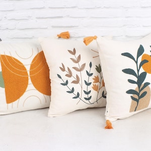 May include: Three square decorative throw pillows with cream-colored fabric and embroidered designs. One pillow features an orange and green geometric design, another has a floral pattern, and the third has a plant and vase motif. Each pillow has an orange tassel.