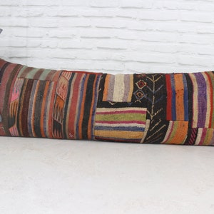 May include: A long, rectangular patchwork cushion with a variety of colours and patterns, including red, orange, yellow, green, blue, purple, and black. The cushion is made of woven fabric and has a fringed edge.