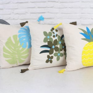 Könnte beinhalten: Drei dekorative Zierkissen mit gestickten Designs. Eines zeigt ein grünes und blaues Blatt, ein anderes grüne und blaue Blumen, und das letzte zeigt eine gelbe Ananas mit blauen und grünen Blättern. Jedes Kissen hat eine Quaste.