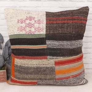 Puede incluir: Un cojín cuadrado de patchwork con una variedad de colores y texturas. El cojín presenta secciones de tela marrón, roja, naranja y crema. Un diseño geométrico rosa es visible en una sección. El cojín es probablemente para la decoración del hogar.