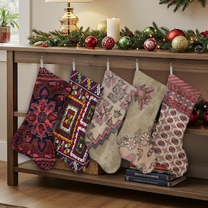 Pu&ograve; includere: Cinque calze di Natale decorate con motivi colorati, rosse, viola, verdi e beige, sono appese a una mensola di legno. Le calze presentano disegni dettagliati. La mensola &egrave; decorata con ornamenti natalizi e verde festivo.