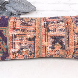 May include: Rectangular decorative pillow with a woven, distressed design. The pillow features a color palette of orange, purple, and navy blue. The pattern includes geometric shapes and vertical stripes, creating a vintage aesthetic. The pillow is placed on a white surface.