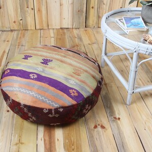May include: A circular floor cushion with a striped design in purple, orange, and beige. The cushion has a dark red border and is on a wooden floor. A white table with a glass top is in the background. The cushion's diameter is approximately 70 cm.