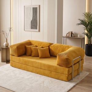 May include: A mustard yellow corduroy sofa with matching throw pillows. The sofa has a low profile and a modern design, with a metal frame. The room features a neutral color palette with a white rug and a framed abstract artwork.