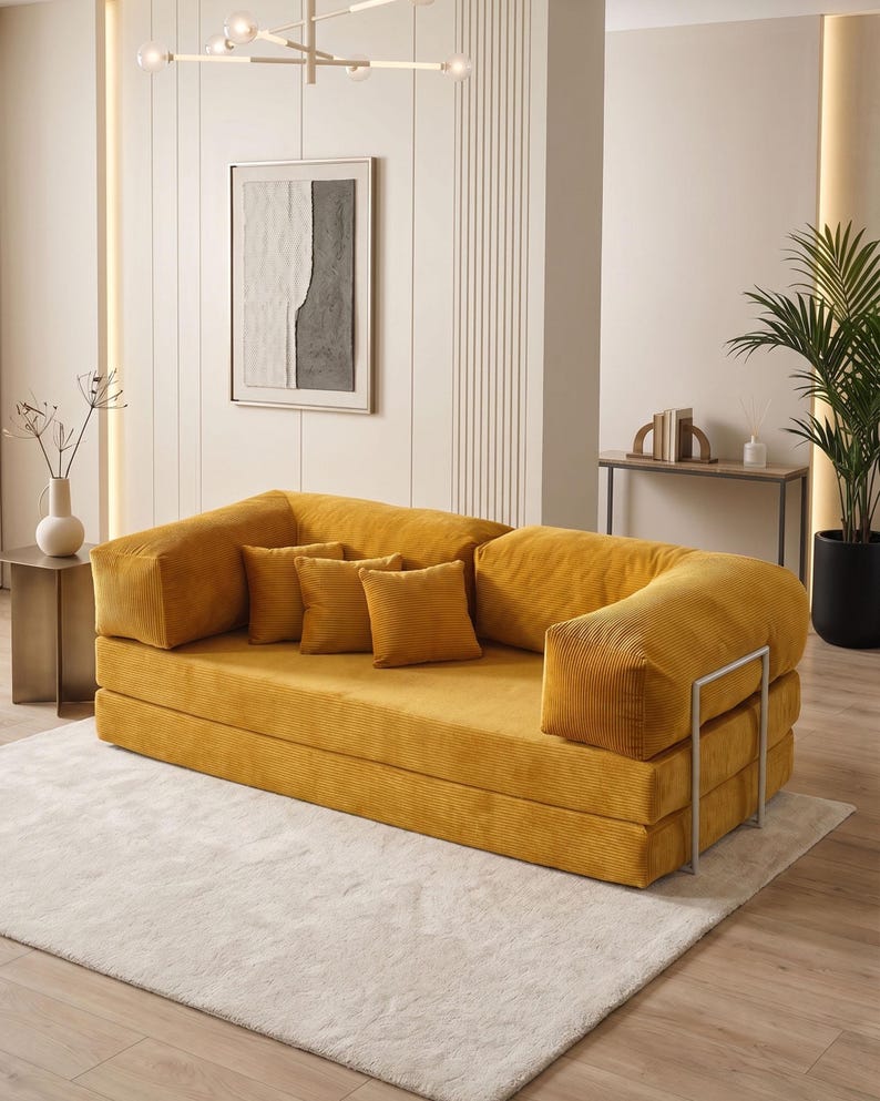 May include: A mustard yellow corduroy sofa with matching throw pillows. The sofa has a low profile and a modern design, with a metal frame. The room features a neutral color palette with a white rug and a framed abstract artwork.