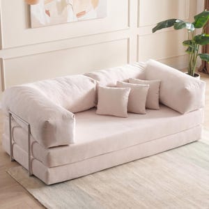 May include: A light pink, corduroy-textured convertible sofa with three matching throw pillows. The sofa has a metal frame and is set on a light-colored rug. The room has a neutral color palette with a painting and a potted plant.